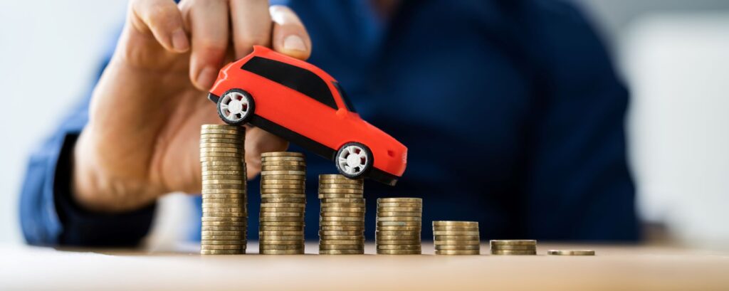 Ein Gutachter in Hanau hebt ein rotes Spielzeugauto auf gestapelten Münzen an, was die Ermittlung des Fahrzeugwerts nach einem Unfall symbolisiert.