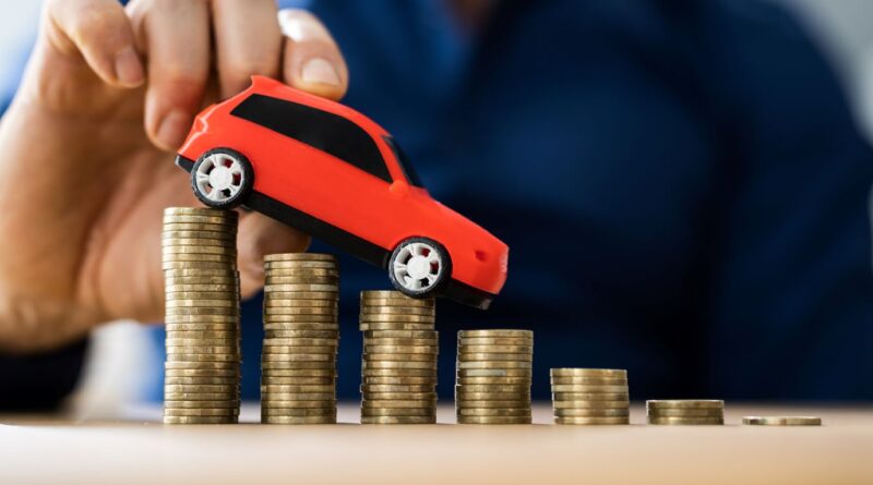 Ein Gutachter in Hanau hebt ein rotes Spielzeugauto auf gestapelten Münzen an, was die Ermittlung des Fahrzeugwerts nach einem Unfall symbolisiert.