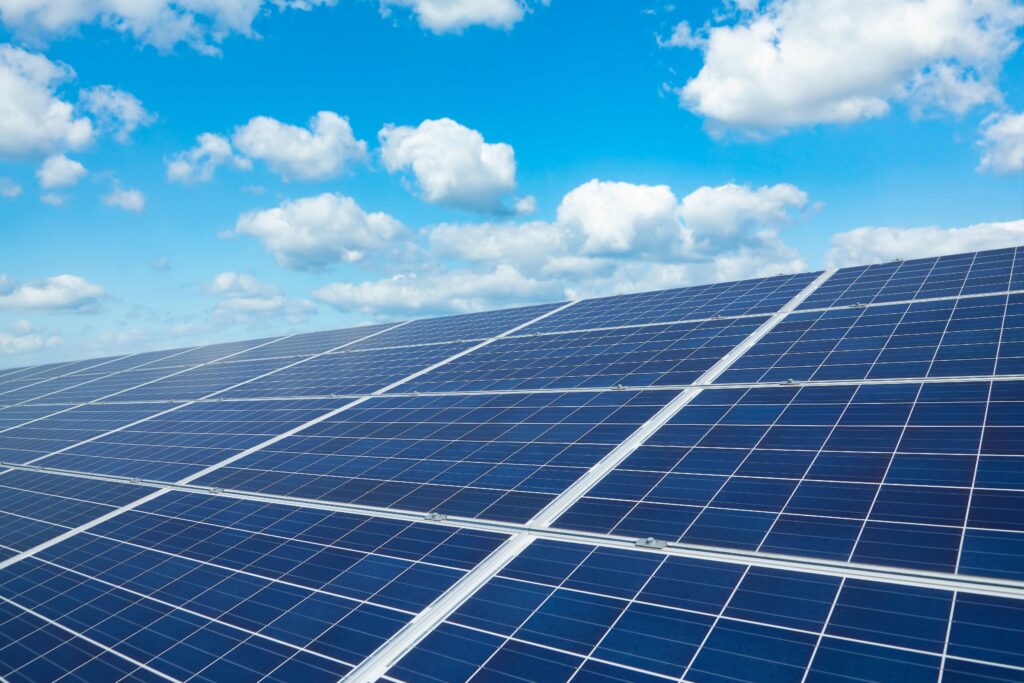 Nahaufnahme einer großen Solaranlage unter einem klaren, blauen Himmel mit vereinzelten weißen Wolken. Die Solarmodule sind gleichmäßig angeordnet und reflektieren das Sonnenlicht.