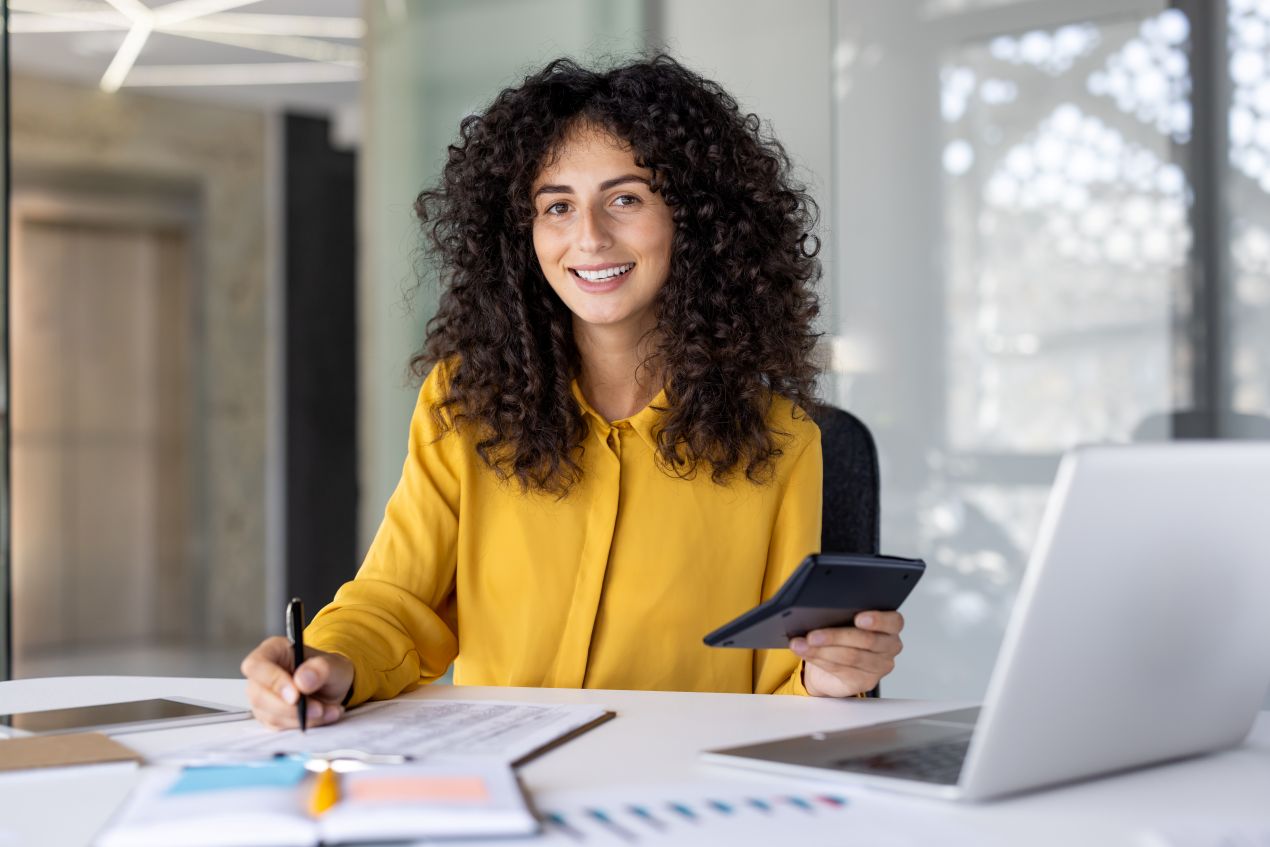 Lächelnde Frau im Büro mit Laptop, Notizen und Taschenrechner – Alternativen zum Masterarbeit schreiben lassen entdecken.