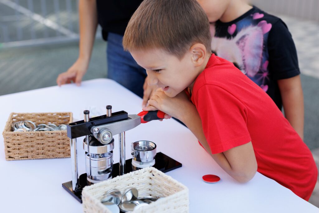 Junge bedient eine manuelle Buttonmaschine an einem Tisch mit Button-Rohlingen